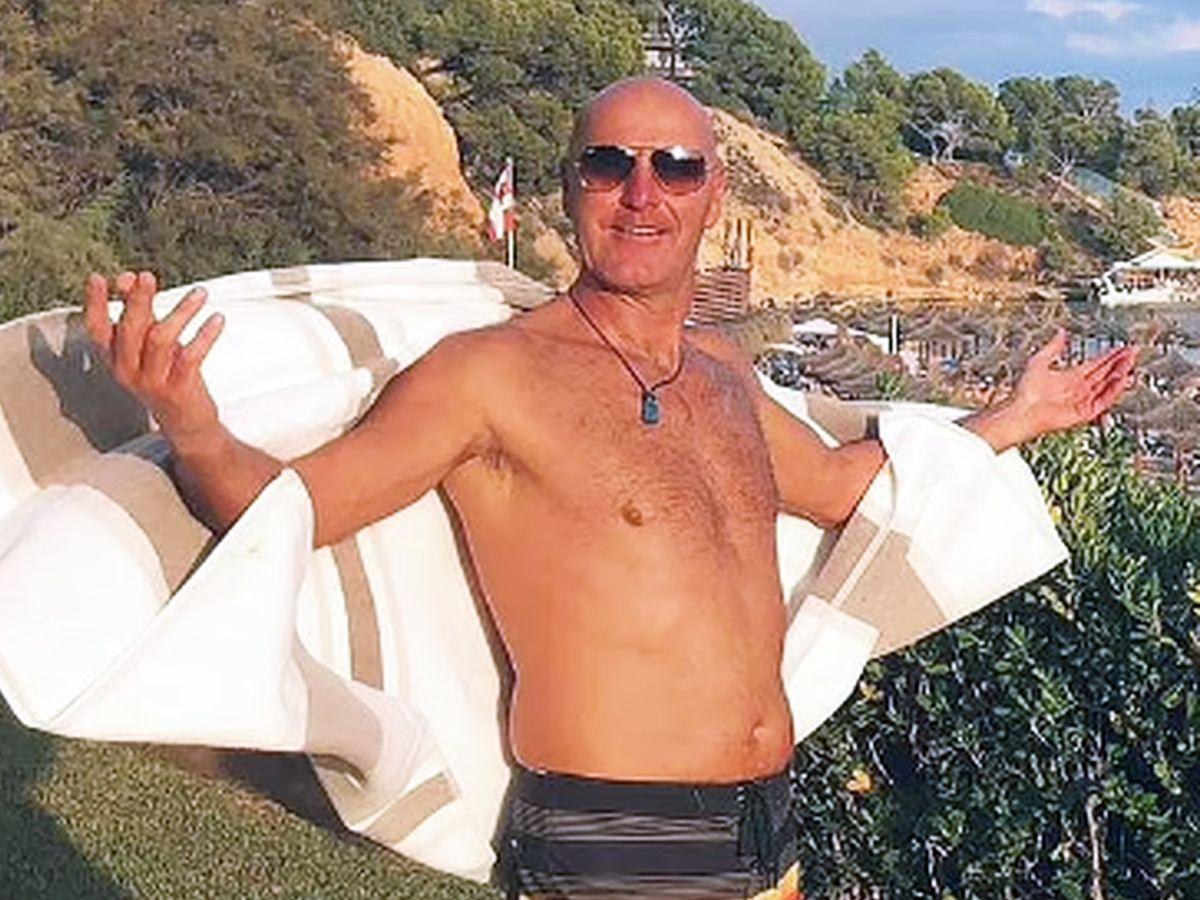 A bald man wearing aviator sunglasses. He has no shirt on and both his arms are splayed out, his lower arms attached to what looks like a white shirt or towel. The sun is shining brightly.