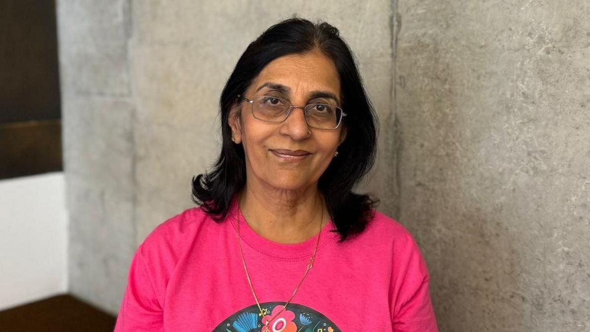 Nisha smiling. She is wearing glasses and a necklace. Her t-shirt is bright pink with flowers on it.