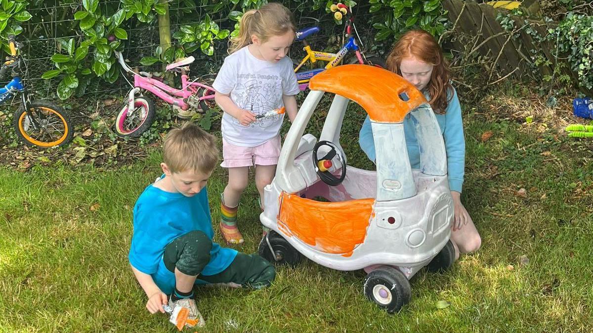 Three kids painting a ride-in toy car in the background bikes and trikes