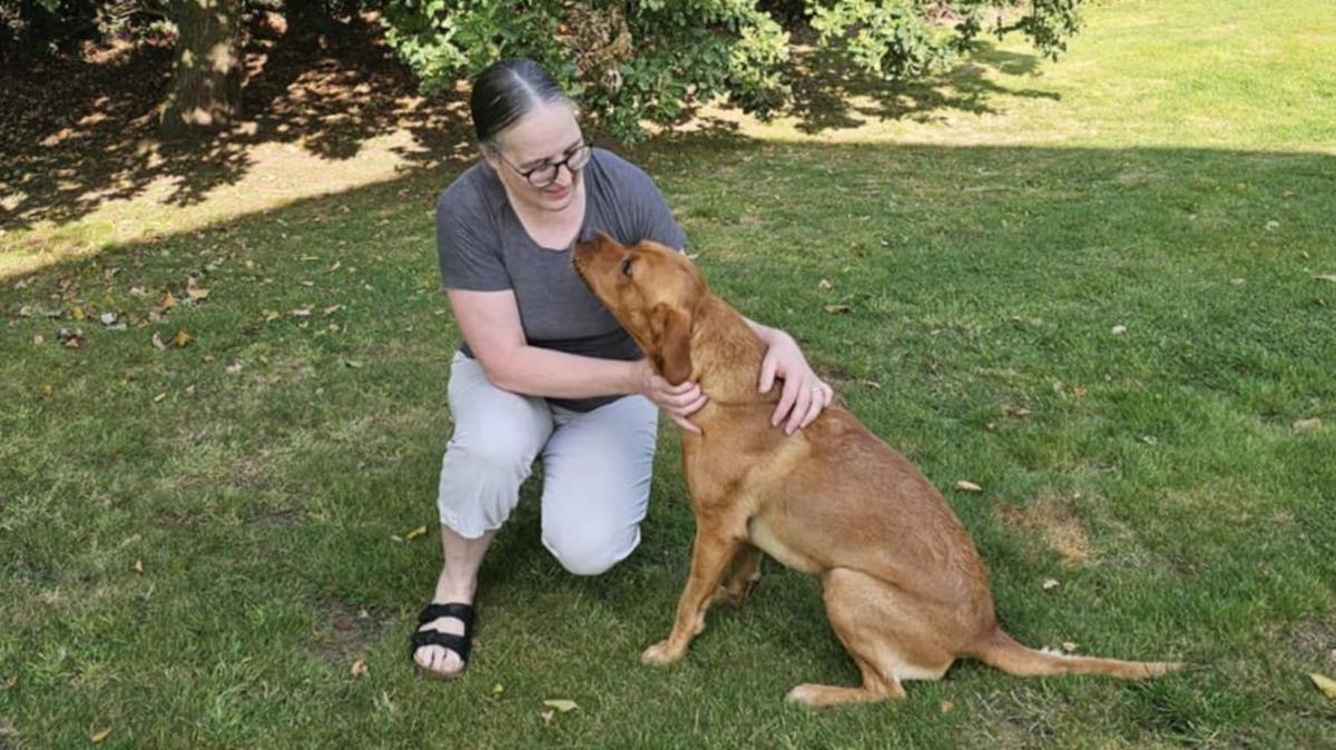 Sam Mawer with her Labrador, Malcolm, in the garden. Malcolm is looking up at Mrs Mawer, who has grey hair and black rimmed glasses.