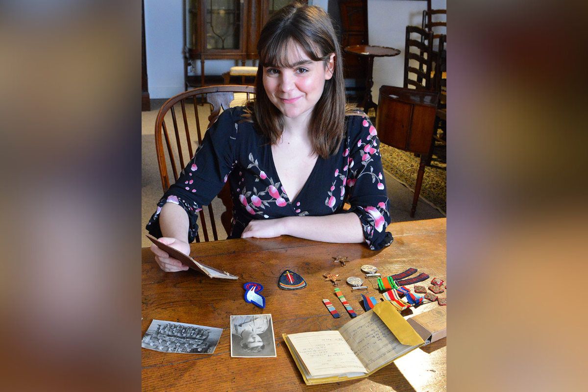 A brunette woman in a floral dress is sat at a wooden table with war memorabilia including photographs, notebooks, and medals spread out in front of her.