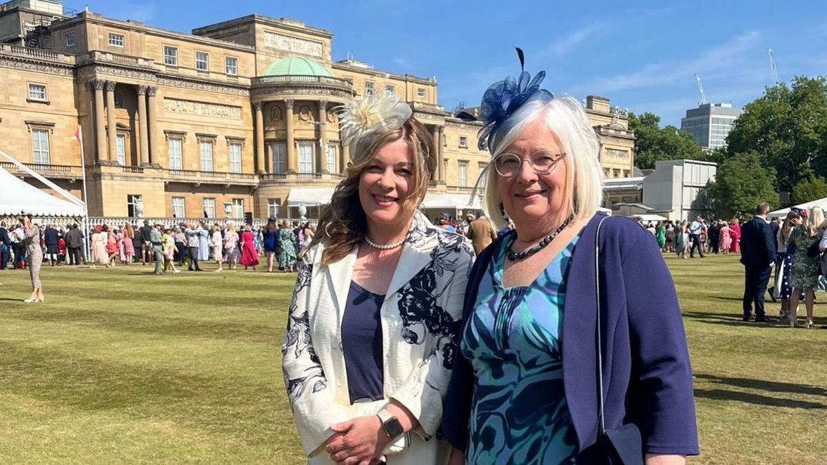 Two ladies are smiling at the camera. They are dressed smartly with fascinators on - the lady on the left in white with a white headpiece, and the lady on the right in blue with a blue headpiece. They are stood in front of a grand building, and in the background there are lots of people stood on the grass.