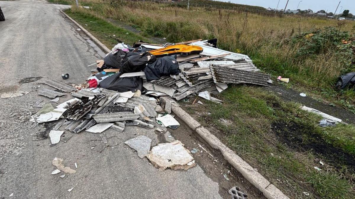 A large pile of fly-tipped waste half on a road and half on a grass verge. The rubbish pile is made up of corrugated roofing sheets and black bags of waste. 