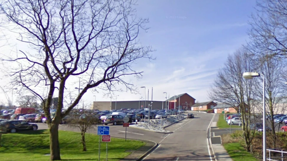 A view of the HMP Lowdham Grange in Nottinghamshire carpark showing a road and cards in the foreground and buildings in the background. 