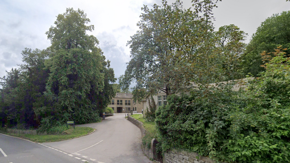 The road entrance to Aldern House is surrounded by trees on one side and a small stone wall and a grass verge on the other. Light brick buildings can be seen in the background at the end of the driveway.