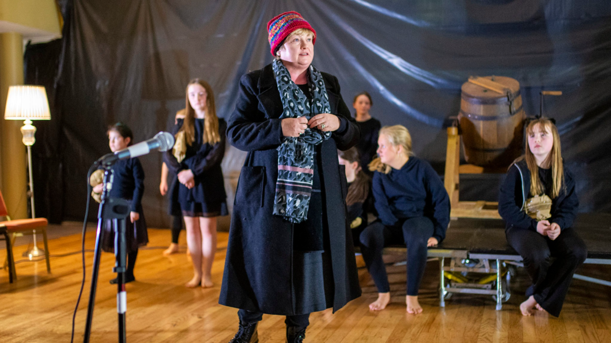 A school play is taking place up at the front is a woman with a hat, black coat and blue patterned scarf. In the background is children wearing a navy top and trousers, or a dress while sitting on a stage.