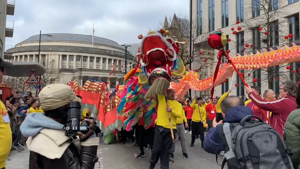 Performers parade a red and golden dragon in front of Manchester's Central Library, down a street lined by crowds, surrounded by press photographers