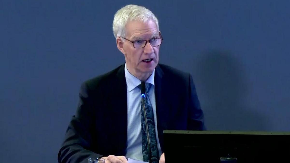 James Baxter at the Omagh Bombing Inquiry. He sits in front of a computer, wearing a blue suit, blue shirt and patterned blue tie. He has glasses and grey hair.