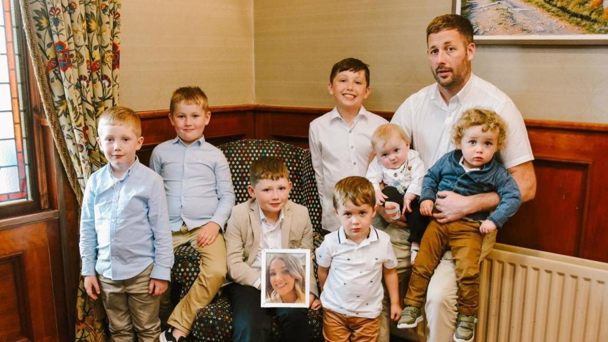 Seven boys all wearing shirts sit on a sofa with their dad and a picture of their mum. 