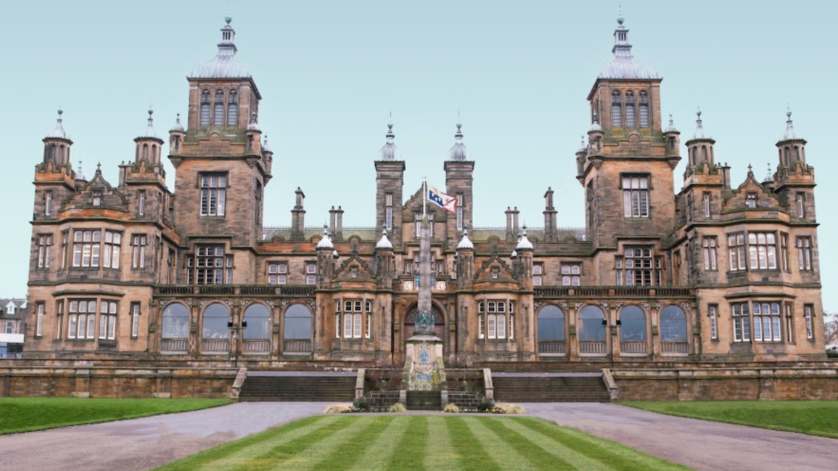 A large historic school building with mutliple towers and turrets, high arched windows, with a flagpole front and centre behind a manicured striped green lawn.