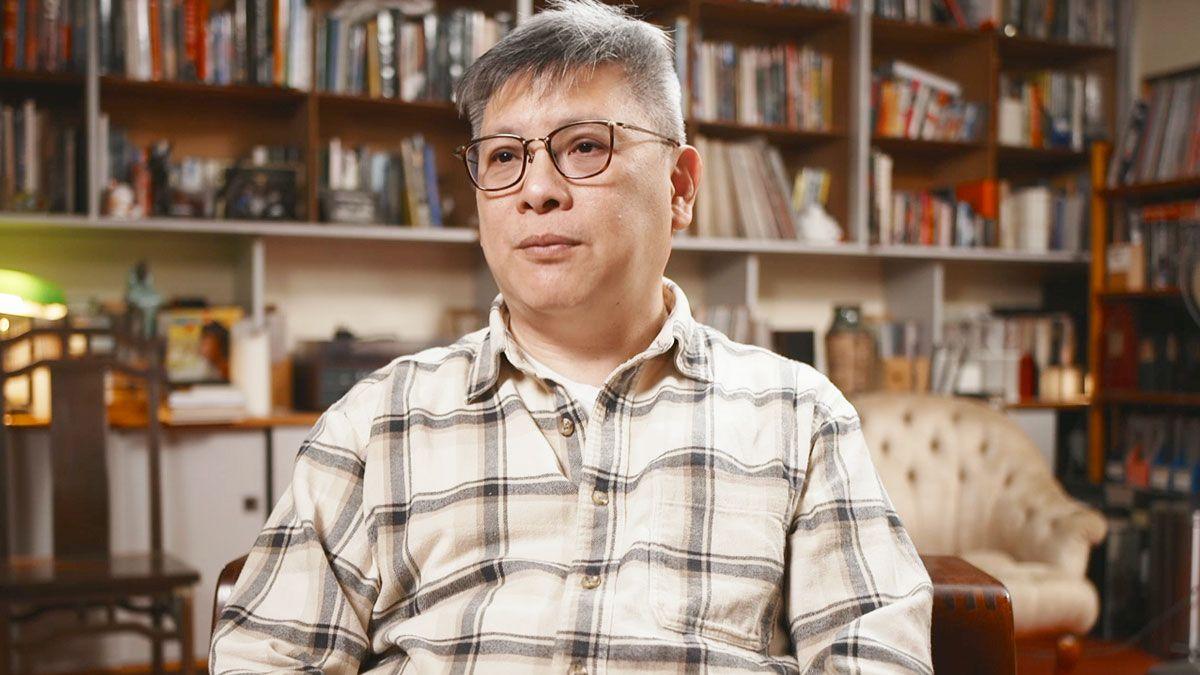 Innes Tang wears glasses and a black and white plaid shirt. He sits in a book-lined room, speaking to the camera