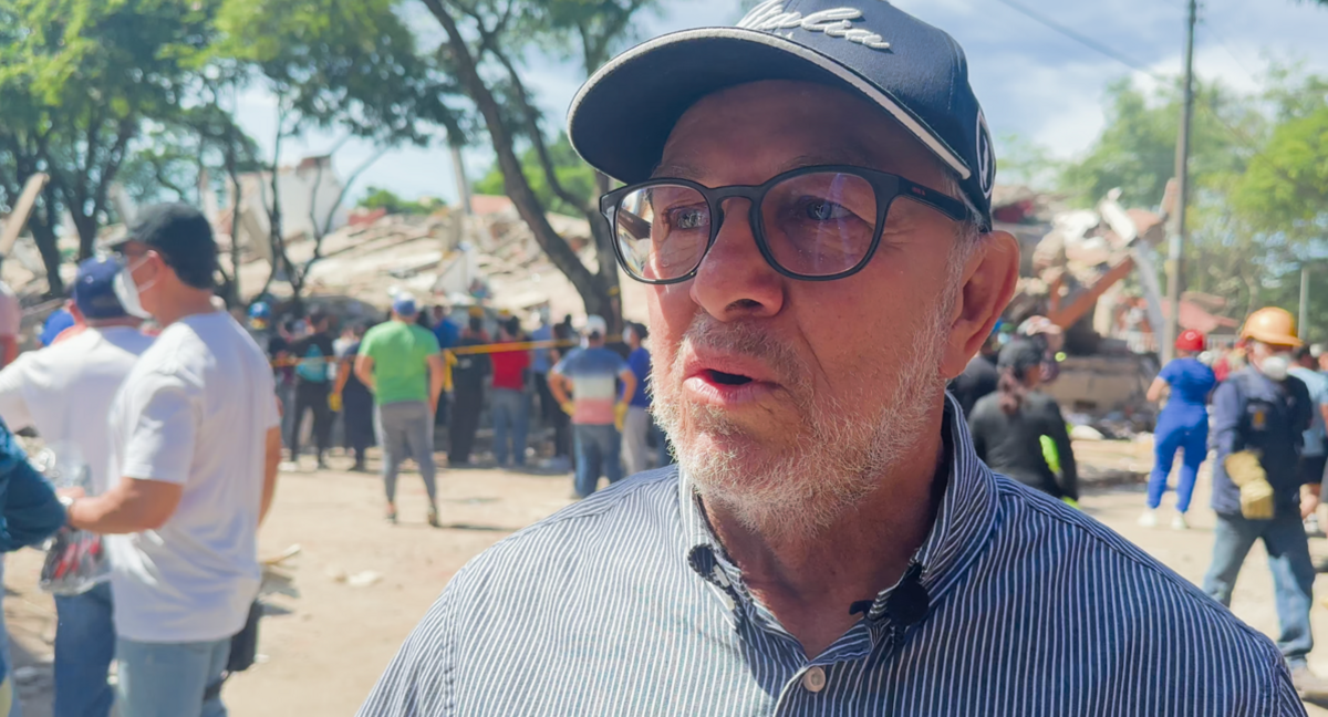 A man with a short white beard, glasses, a baseball cap, and a buttoned up shirt with an oxford collar 