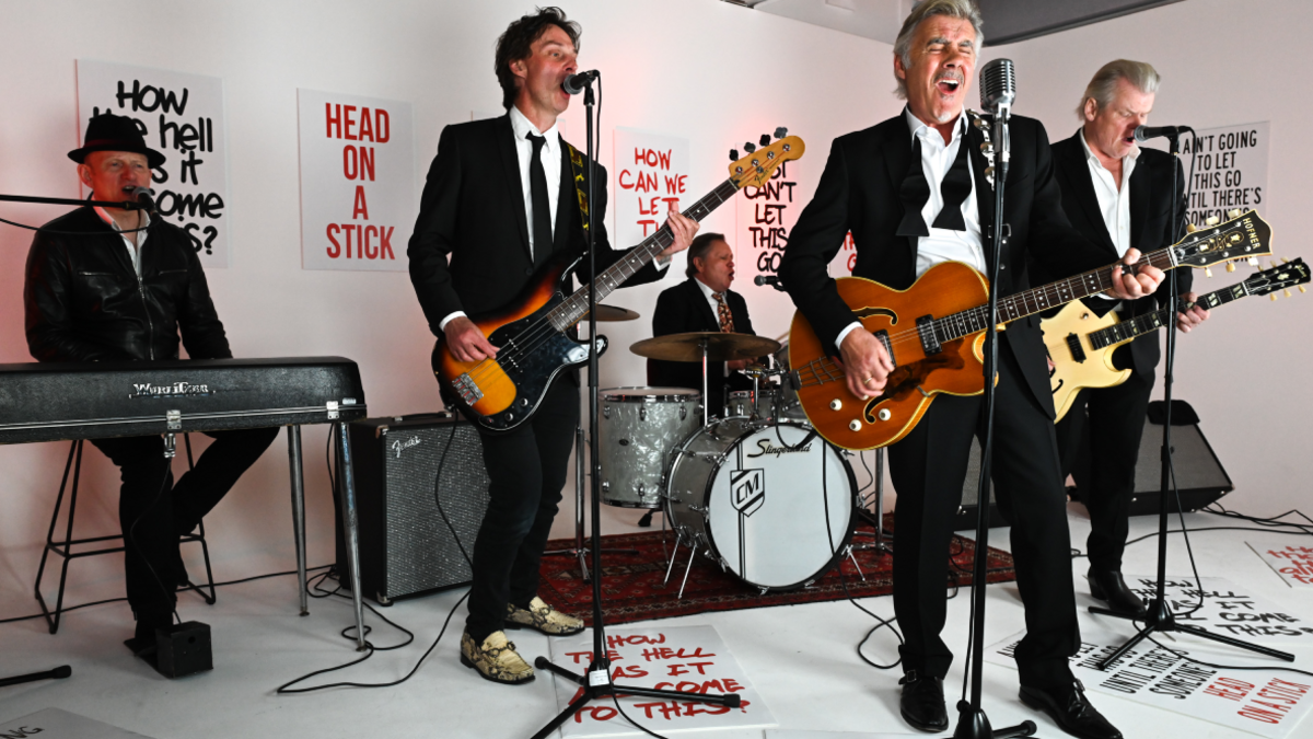 Glen Matlock, playing a guitar and singing, performing with the four other members of his band, who are on keyboards, bass, guitar and drums. All are dressed in black and white, and there are slogans written on the wall and on the floor, such as 'head on a stick'