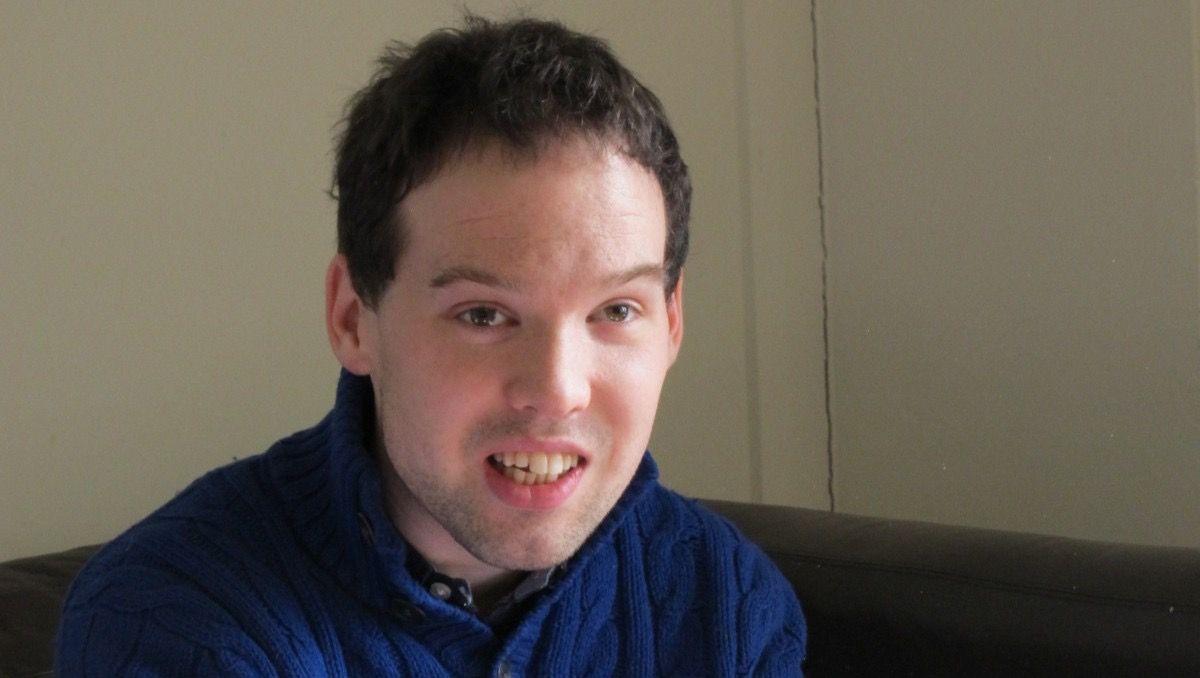 Tom Wentworth is pictured in a blue knitted jumper sitting indoors on a dark sofa, facing the camera. The image is a close-up portrait with a plain cream-coloured wall in the background and soft natural lighting.