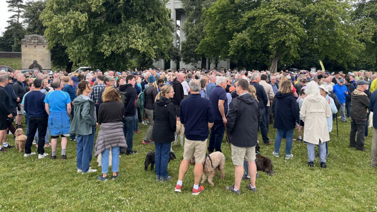 A crowd of people gather on the Downs near the water tower - they mostly wear waterproof coats and there are some dogs amongst them