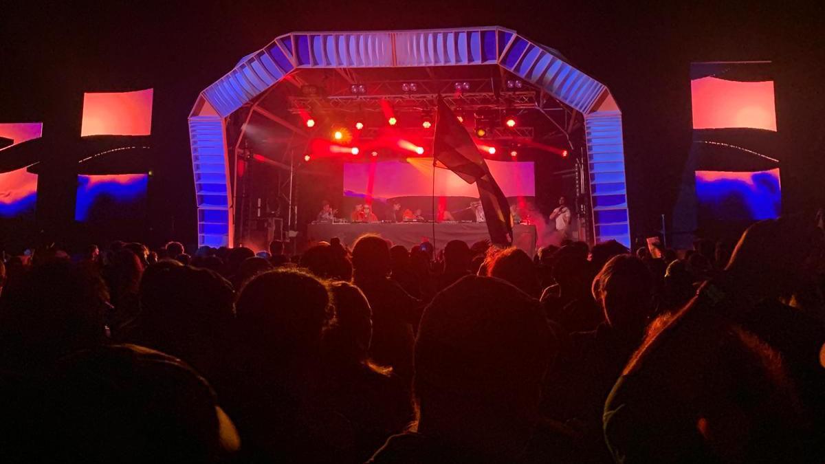 A crowd of people face an outdoor music stage at a festival. The sky is dark and the stage is lit in red, pink and blue. 