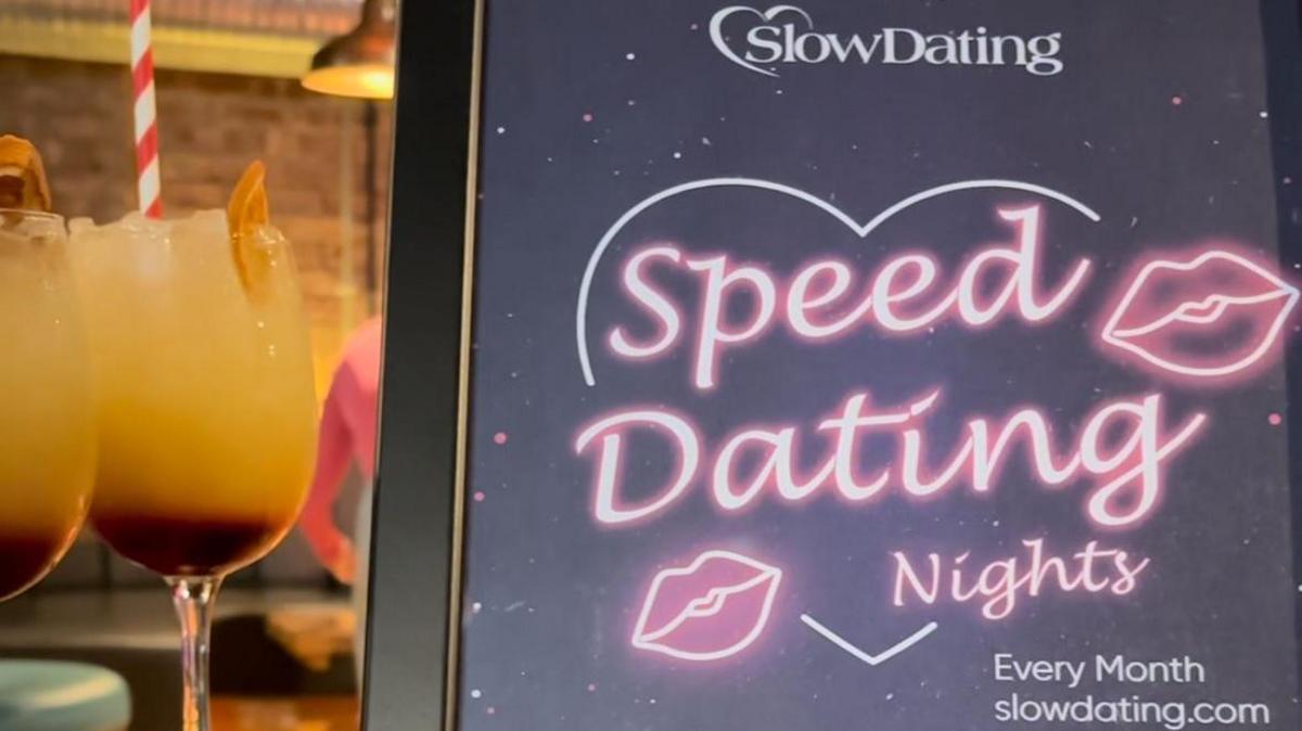 Poster advertising Speed Dating Nights. The text is pink, written inside a heart. There are lip prints next to the text. Drinks can be seen in the background of the photo. 