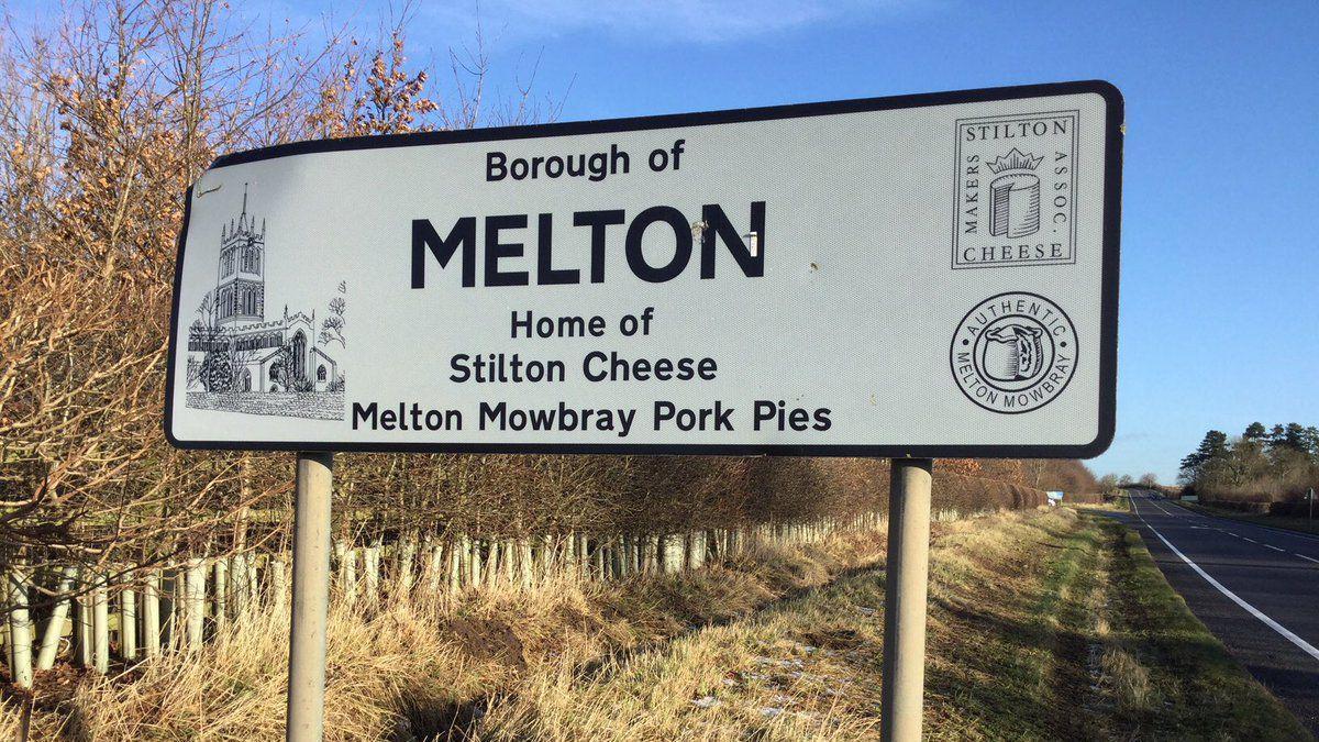 A white road sign with black lettering saying "Borough of Melton. Home of Stilton Cheese Melton Mowbray Pork Pies"