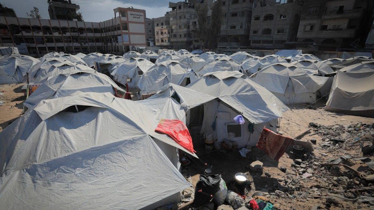 Lots of grey tents to provide shelter to displaced families line a dusty square of land in the middle of damaged buildings. 