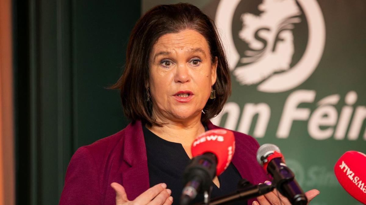 Mary Lou McDonald, a woman with shoulder-length brown hair, speaks at a row of press microphones with the Sinn Fein logo on the backdrop behind her