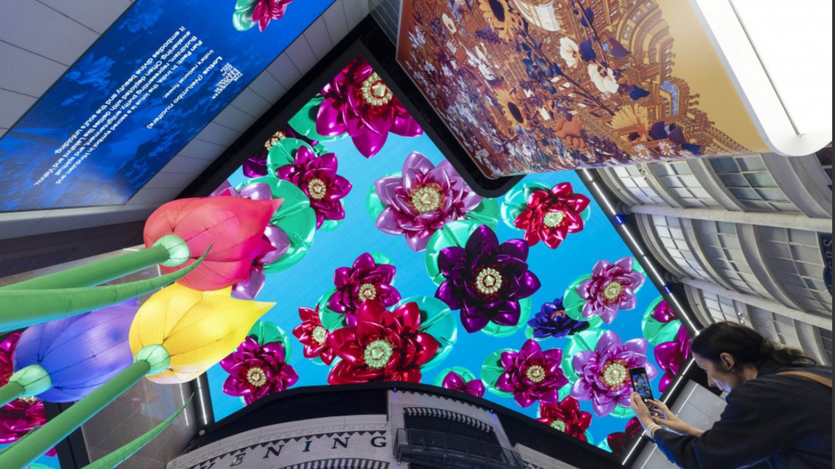 A man takes a photo of a digital image of red and purple flowers on a ceiling.