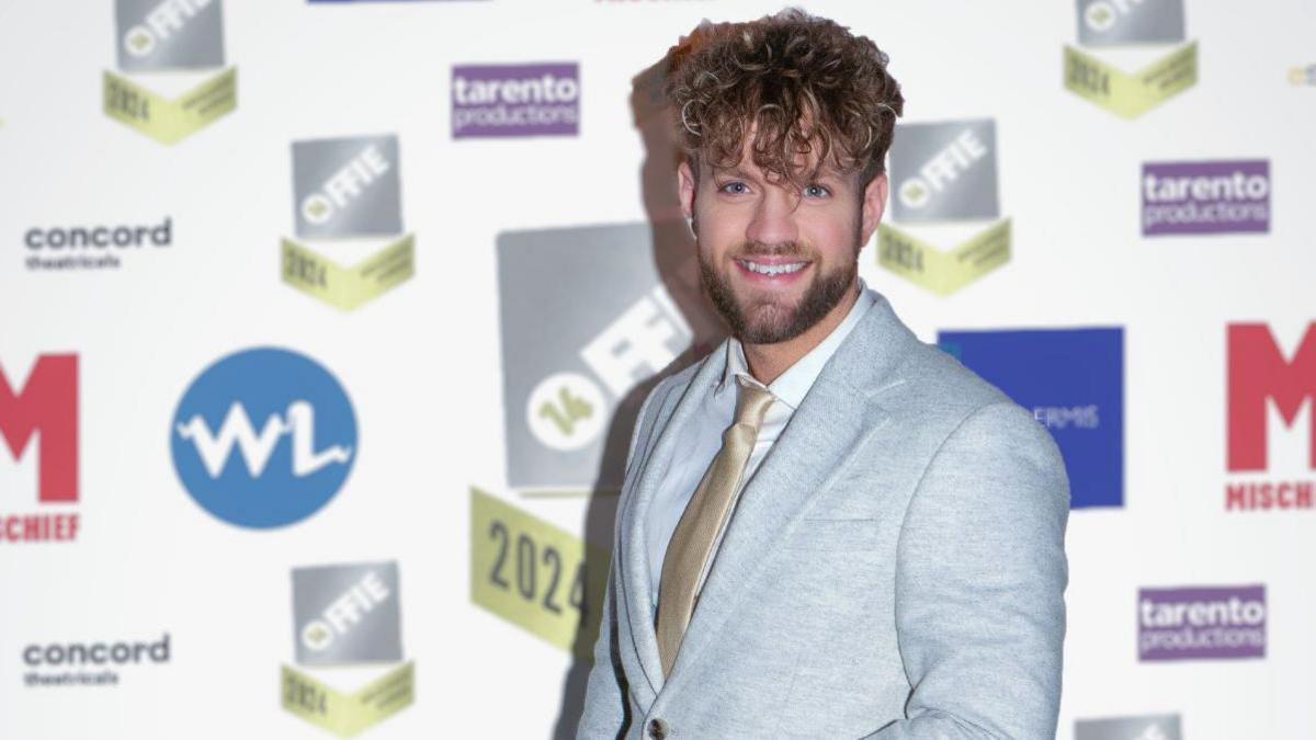 A smiling man with fair curly hair and a beard standing in front of an awards show board covered in logos. He is wearing a light grey suit jacket, gold tie and white shirt.