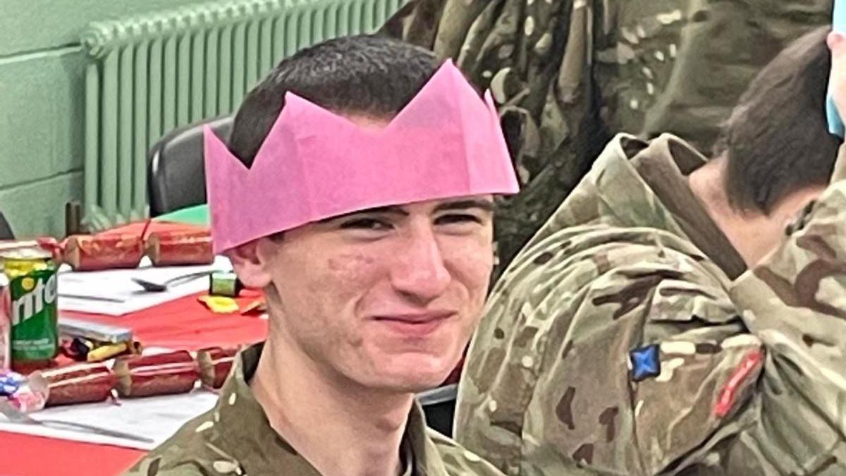 Logan MacPhail. A young man with dark hair and wearing army camouflage clothing smiles at the camera. He is wearing a pink paper crown from a Christmas cracker.