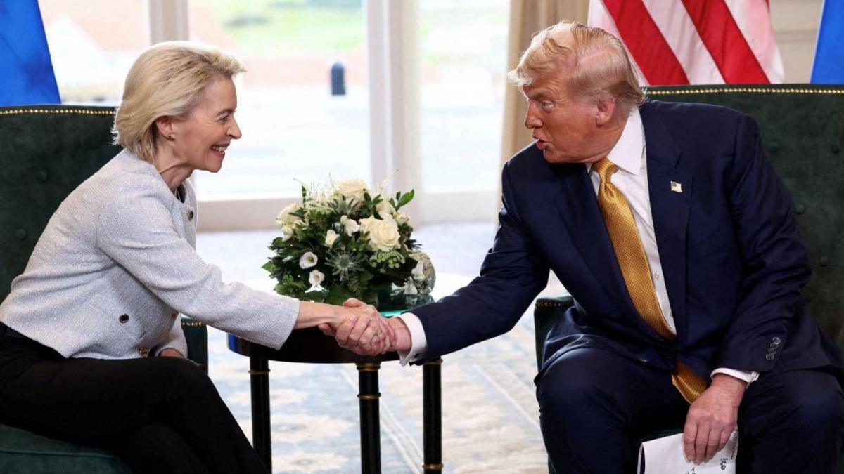 Ursula von der Leyen shaking hands with Donald Trump. They are both sitting on green chairs. Von der Leyen is wearing a grey blazer and black trousers. Trump is wearing a navy suit, white shirt and gold tie.
