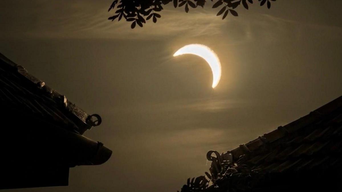 The semi-circled sun in a dark sky viewed between two rooftops.