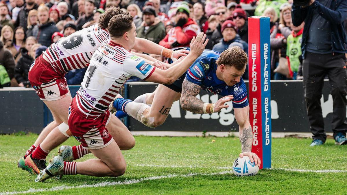 Wakefield's Tom Johnstone diving towards the line to score a try one-handed for his side under pressure from two Wigan defenders in the corner.
