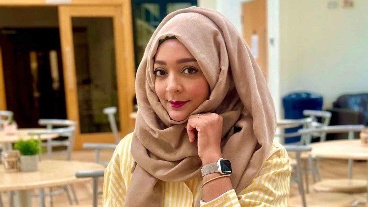 A woman wearing a brown hijab and yellow and white striped top smiling, she has her chin resting on her hand and she has a watch on her wrist. 