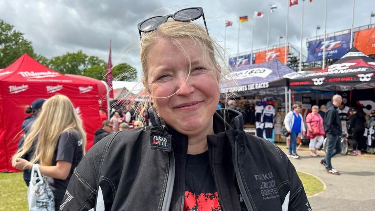 Sabine Wellbrock has blond hair tied up, wearing a black jacket and sunglasses on her head. There are crowds walking behind at the TT Grandstand.