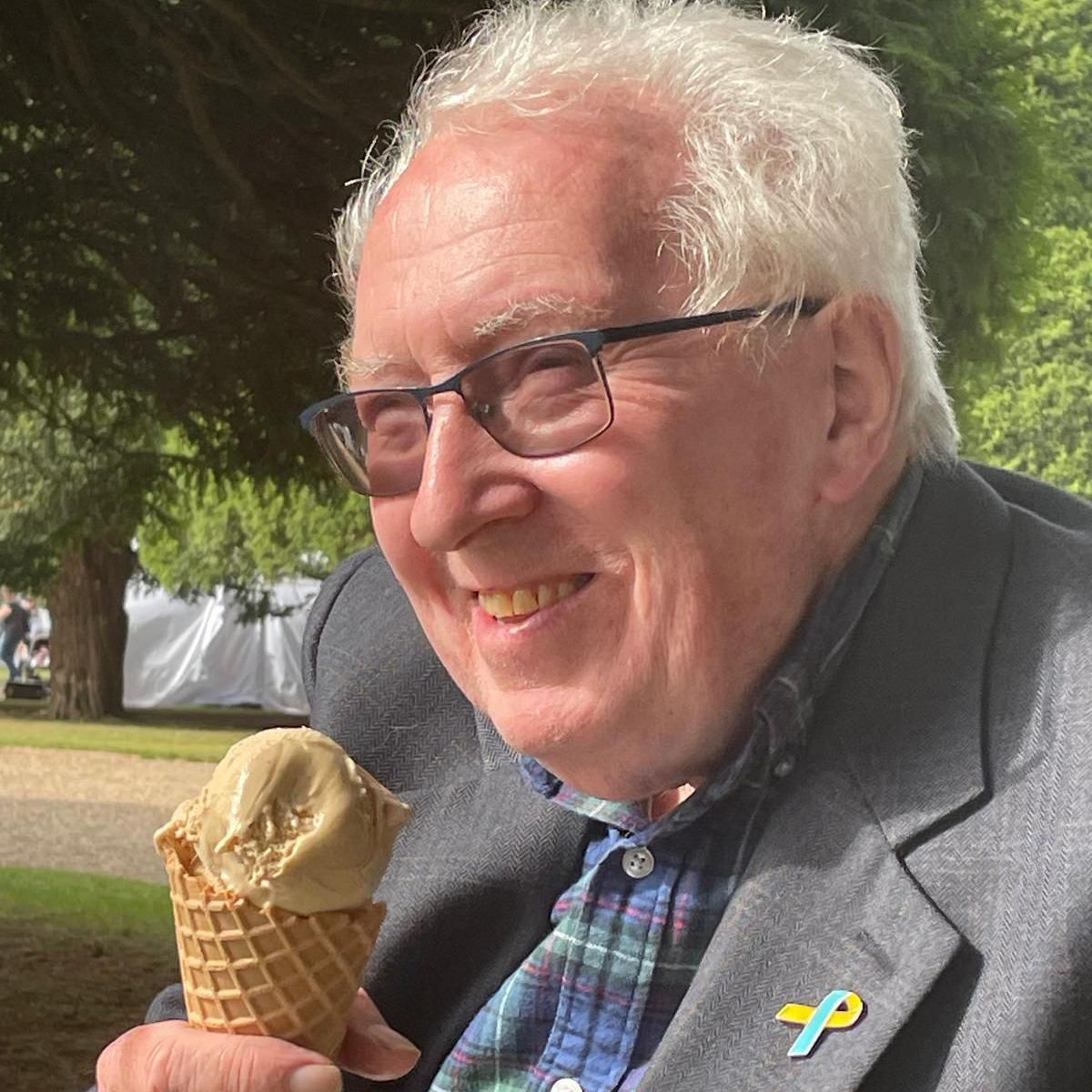 Jo Quigley smiling with an ice cream. He is sitting in parkland and wearing a grey jacket and blue-checked shirt