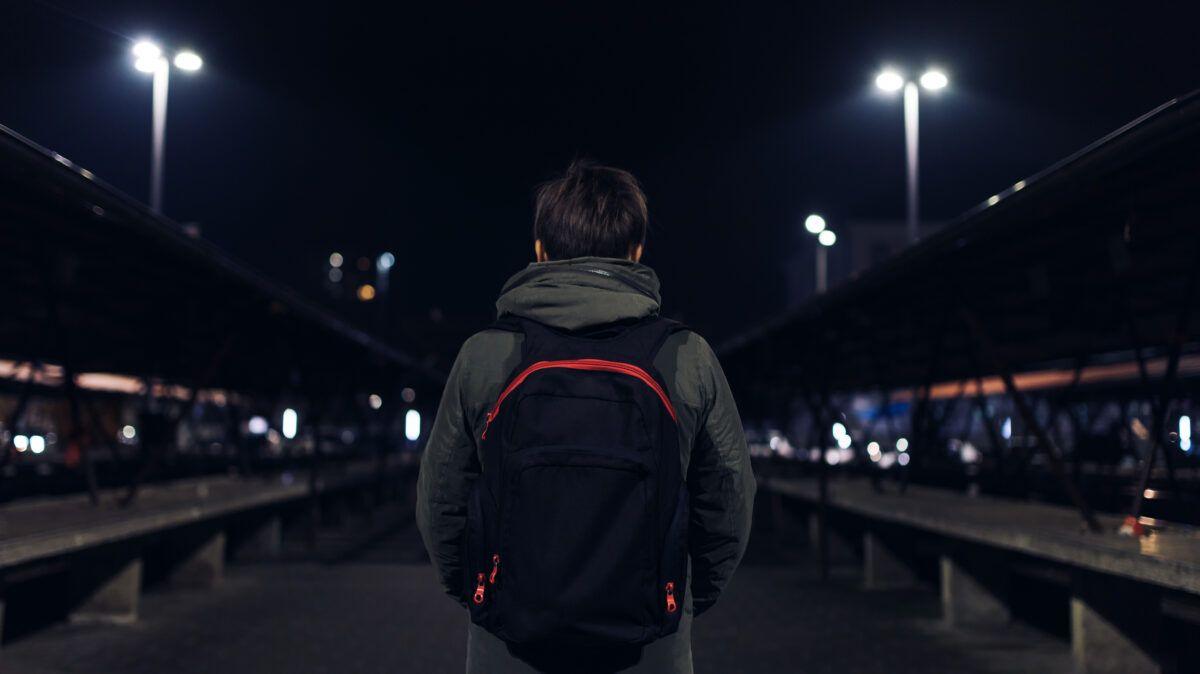 A boy with his back to the camera wearing a backpack. It is night time and he appears to be at a train station.