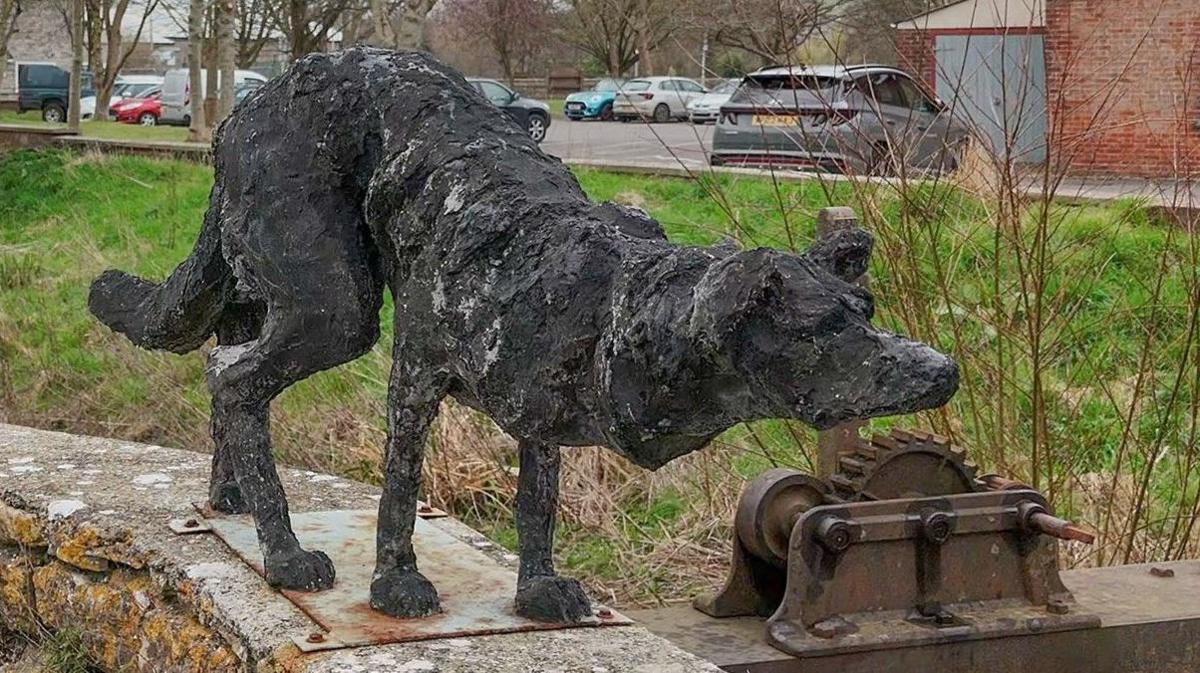 The black cast dog sculpture standing on top off a wall with the weir mechanics in the background. The dog resembles a border collie when it stalks sheep. Its head is low, tail is down and ears are forward.