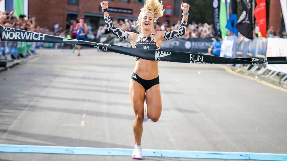 Holly Archer, who has blonde hair and is wearing black running pants and black and white arm warmers, crossing the finish line with spectators in the background. She is holding her arms above her head as she celebrates.