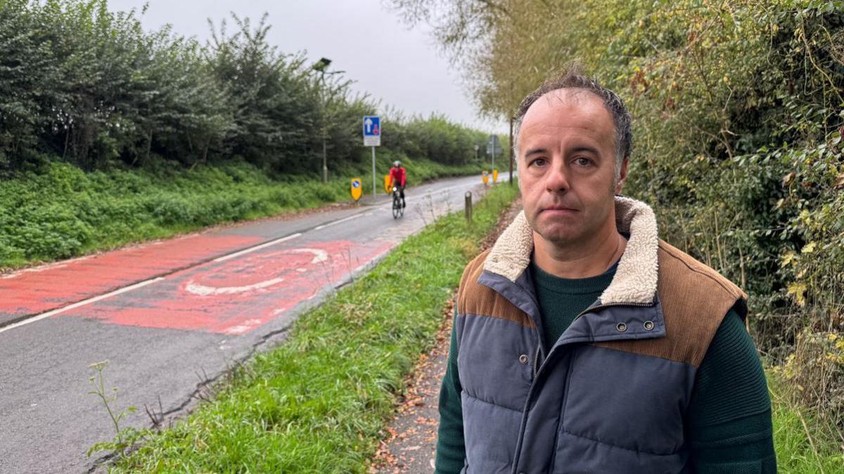 Paul Hayward is standing on the side of Drayton Road. He is looking at the camera. A cyclists is approaching on the road behind him. It is an overcast day.