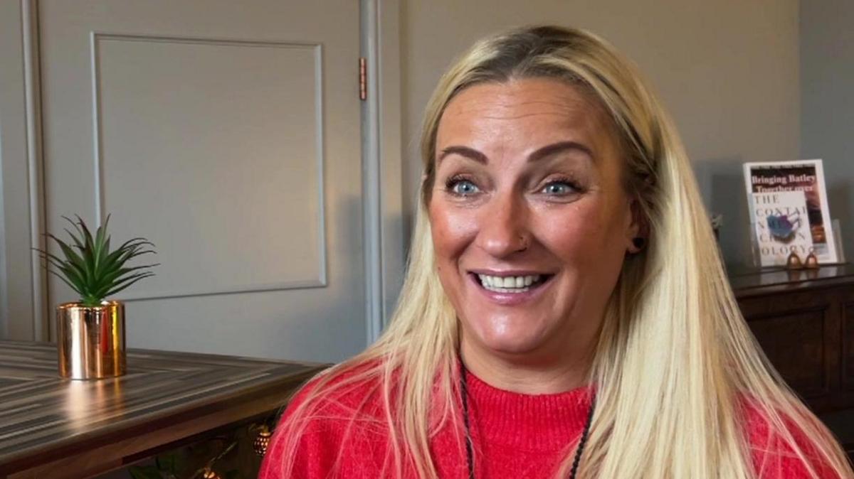 A lady with long blonde hair with a red jumper on is sat next to a bar area. There is a plant on the bar.