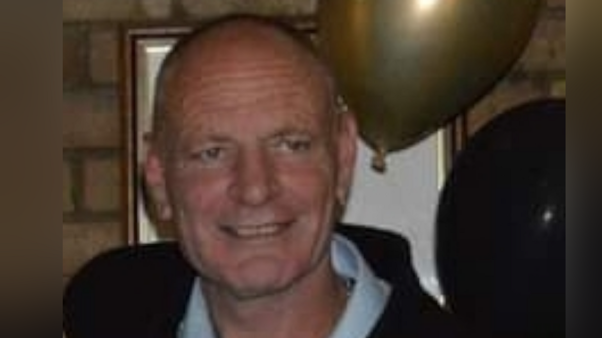 Close up of Gary Hindle in a white shirt and dark jacket. He is standing in front of a picture frame and has a gold and a black balloon by his head.