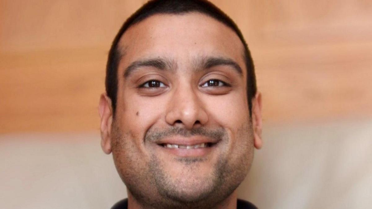 Neil Lakhani is smiling towards the camera. He has brown eyes, dark, cropped, hair and stubble.