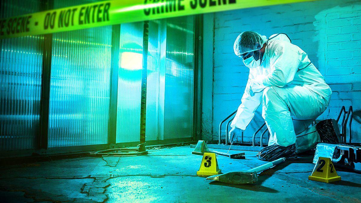 a man in white coveralls, a plastic hair cap and a face mask swabs a plank of wood with a cotton bud. he is at a crime scene, and there is crime scene do not enter tape in front of him