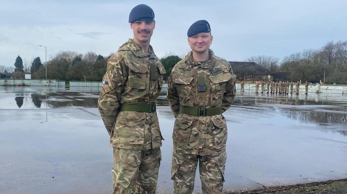 Two men standing with their hands behind their backs, in army camouflage. 
They are wearing blue hats and have a green belt around their waist