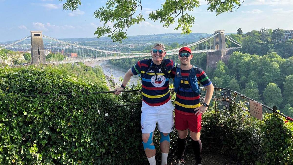 Simon Pilkington, pictured right, stands next to Rob Dowling, pictured left, in front of the Clifton Suspension Bridge. They are both wearing tops with black, red, yellow, blue and green stripes.