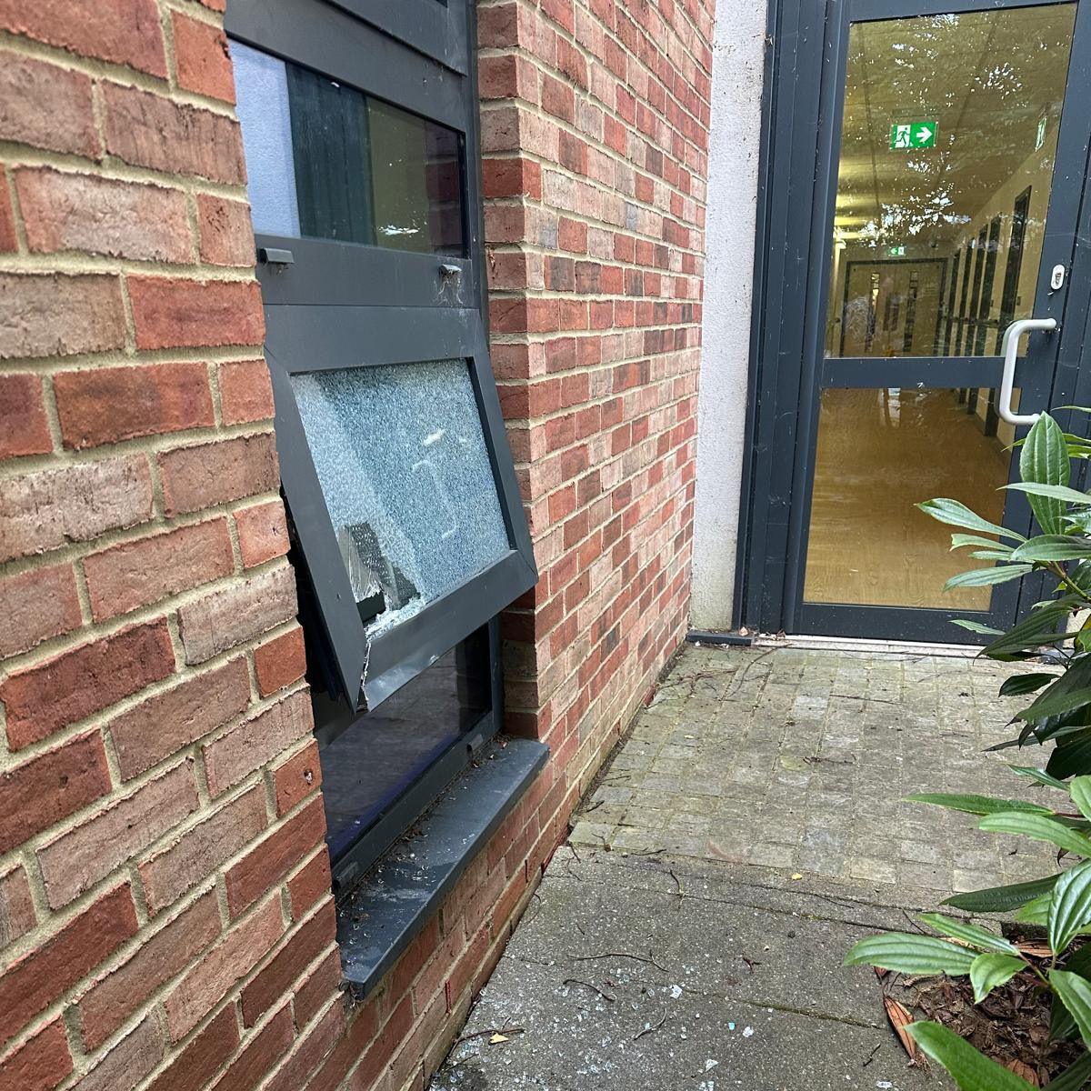 The broken window which was smashed by thieves. The glass is shattered and the frame has been broken. Glass lies on the concrete paving underneath the window.