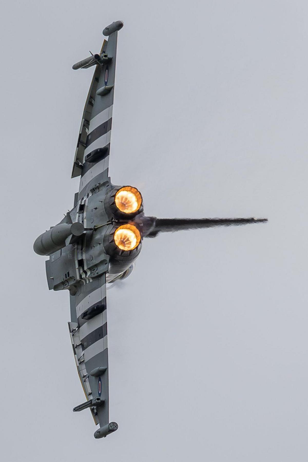 A streamlined plane flies through the sky on its side. Its afterburners are on and glowing bright orange.