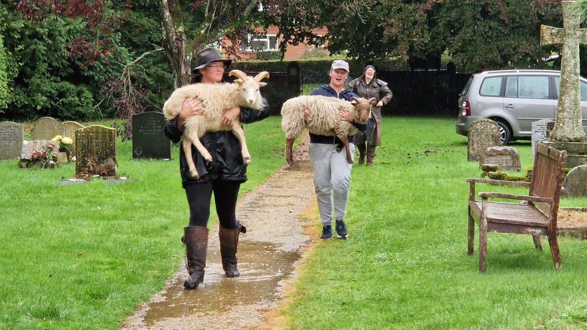 a church graveyard with three people wearing rain gear and carrying the sheep down the path when they first arrived at the church