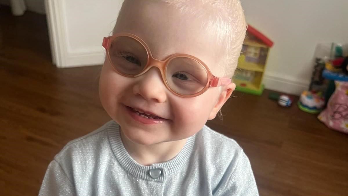 Raya has light blonde hair and is wearing orange glasses and a grey cardigan. She's smiling at the camera and there's a wooden floor, a white wall and some children's toys behind her.