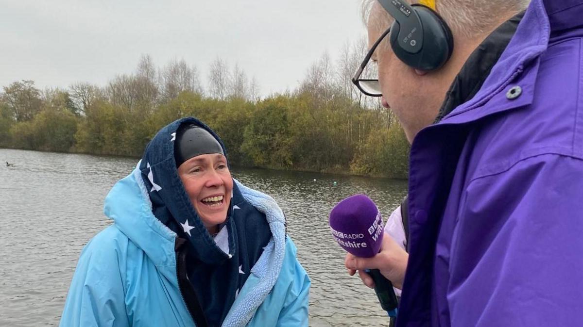 Reporter Kelly Morgan standing next to a lake wearing a lot of layers, including a blue coat. She is being interviewed by a man in a priple jacket holding a purple Radio Wiltshire microphone.