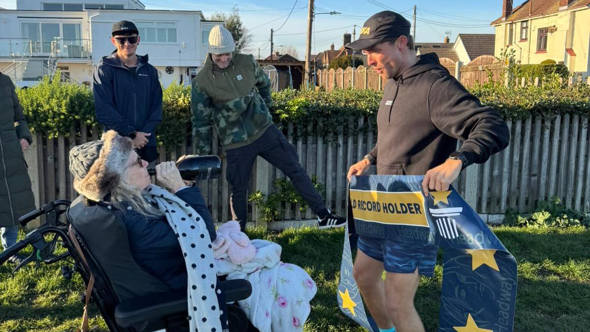 A man in running gear holding a blue banner that says "world record holder". He is standing in front of a woman who is drinking from a bottle of wine. Two men are smiling behind them.