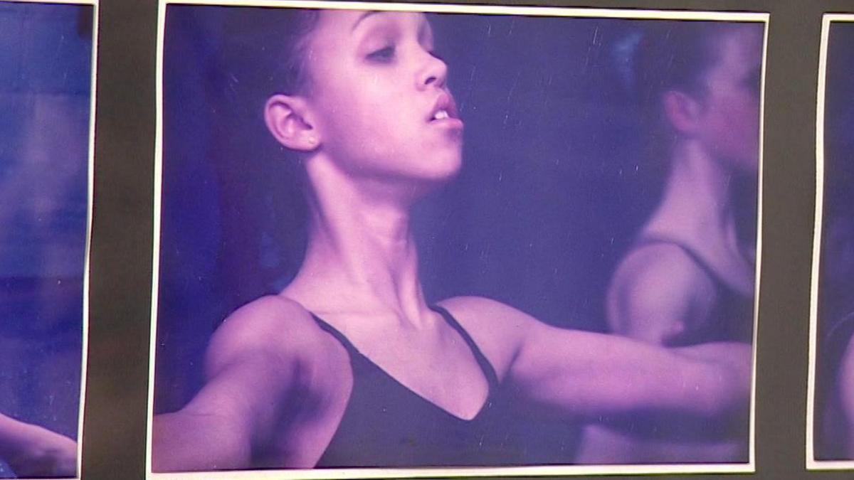 A black display board holding a photo of 12-year-old girl wearing a plain black leotard as she holds her arms out in dance, her mouth slightly open. Her hair is tied back as she performs. Another dancer can be seen beside her.
